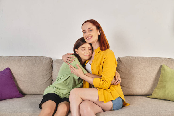 cheerful woman with teenage daughter sitting and embracing on cozy couch in modern living room - Photo, Image