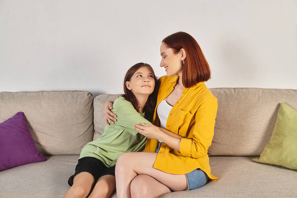 mujer alegre con hija adolescente abrazándose y mirándose en el sofá acogedor en la sala de estar - Foto, Imagen