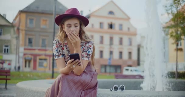 Gyönyörű szőke nő kalapot használ okos telefon ül ellen szökőkút a parkban - Felvétel, videó