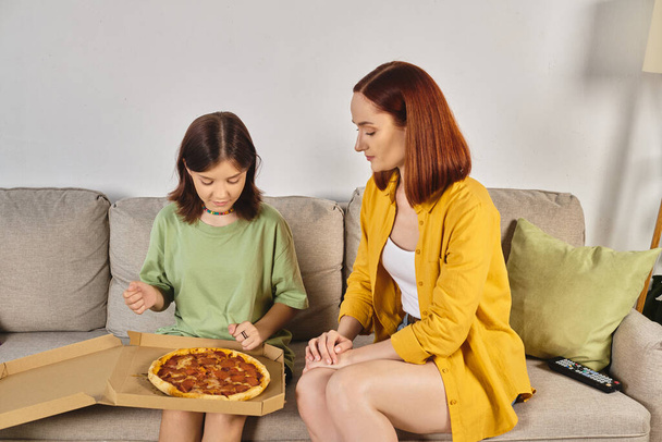 preteen κορίτσι κάθεται στον καναπέ με νόστιμα πίτσα κοντά χαμογελαστή μητέρα στον καναπέ στο σαλόνι - Φωτογραφία, εικόνα