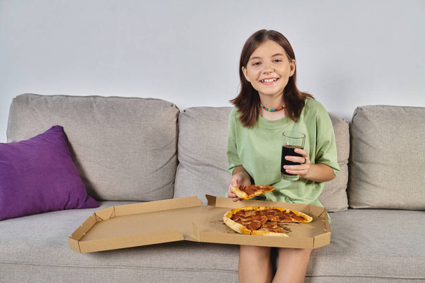 radosna nastolatka patrząc w kamerę siedząc na kanapie z pizzą i sodą, czas posiłku - Zdjęcie, obraz