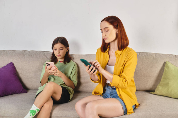 Erwachsene Frau mit Teenager-Tochter nutzt Smartphone, während sie im modernen Wohnzimmer auf der Couch sitzt - Foto, Bild
