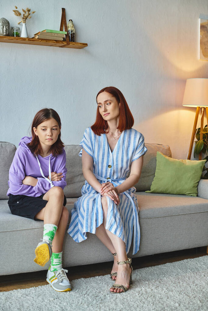 thoughtful woman looking at frustrated teenage daughter on couch in living room, care and support - Photo, Image