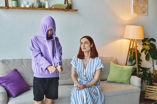 worried and upset woman talking to offended daughter hiding in hood in living room, generation gap - Photo, Image
