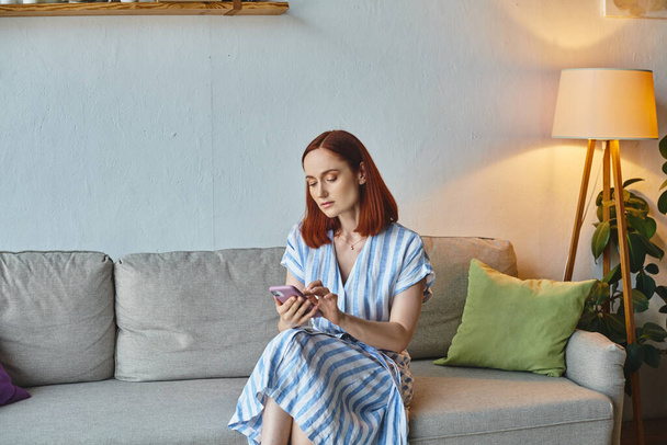 felnőtt nő üzenetküldés mobiltelefonon, miközben ül a kényelmes kanapén a nappaliban, családi élet - Fotó, kép