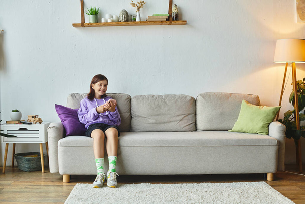 alegre adolescente navegar por las redes sociales mientras está sentado en cómodo sofá en la sala de estar moderna - Foto, imagen