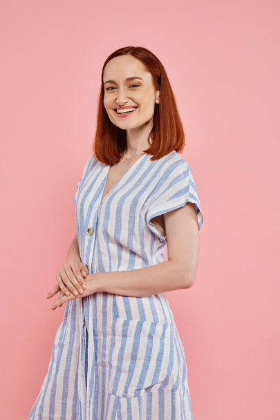 joyful and trendy redhead woman in striped dress looking at camera on pink backdrop, studio portrait - 写真・画像