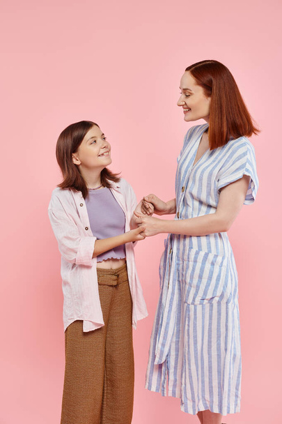 radosna i modna kobieta z nastoletnią córką trzymającą się za ręce i uśmiechającą się do siebie na różowo - Zdjęcie, obraz