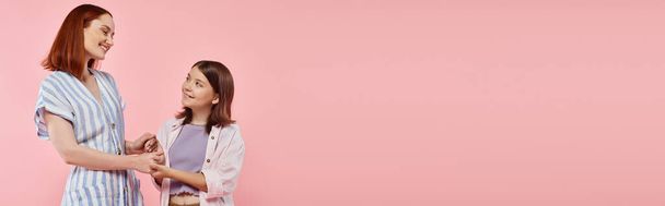 cheerful and trendy woman with teen daughter holding hands and smiling at each other on pink, banner - Photo, Image