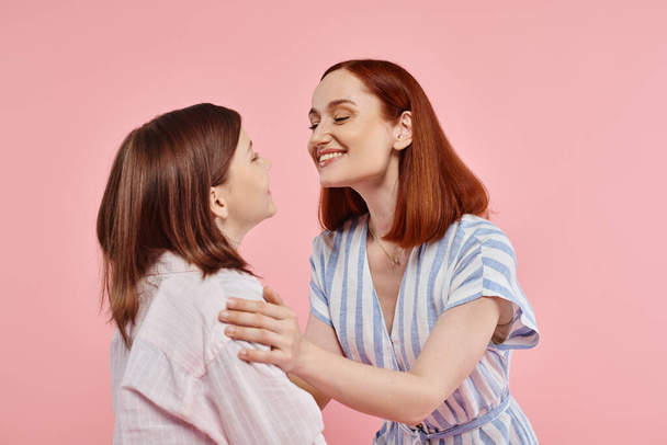 joyful and stylish woman with teenage daughter smiling at each other on pink backdrop in studio - Photo, Image