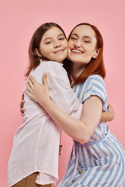 mujer alegre con hija adolescente en elegante atuendo casual abrazando y mirando a la cámara en rosa - Foto, imagen