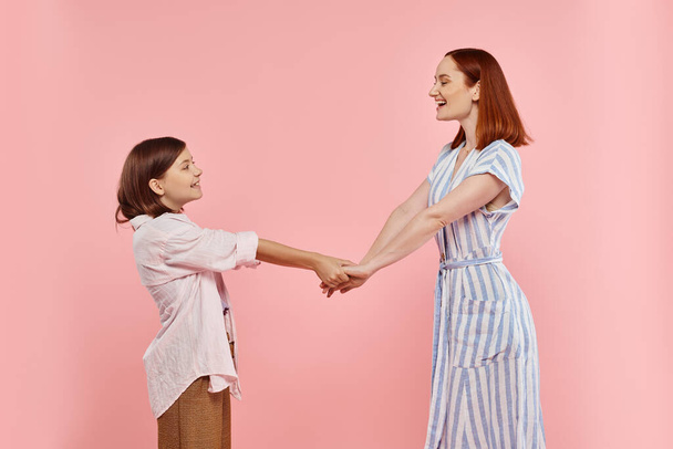 vista lateral de mujer alegre y de moda con hija adolescente cogida de la mano en rosa, familia feliz - Foto, imagen