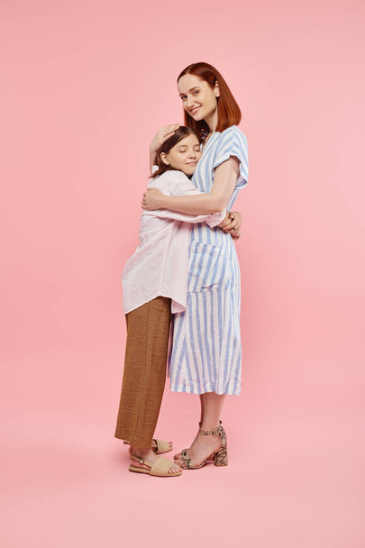 χαρούμενη μοντέρνα γυναίκα αγκαλιάζει έφηβη κόρη και χαμογελά στην κάμερα σε ροζ φόντο, πλήρους μήκους - Φωτογραφία, εικόνα