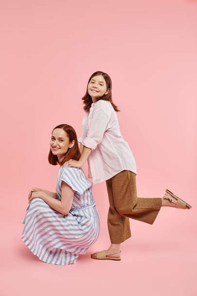 cheerful stylish woman sitting on haunches near teen daughter posing on pink backdrop, fun and joy - Photo, Image