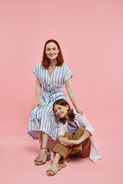 happy and stylish woman with teenage daughter sitting and looking at camera on pink backdrop - Photo, Image