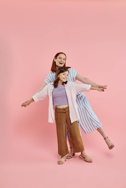 cheerful and stylish woman holding hands of preteen daughter while posing on pink backdrop - Photo, Image