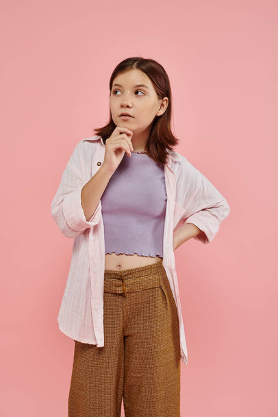 menina adolescente pensativa e elegante de pé com a mão no quadril e olhando para o fundo rosa - Foto, Imagem