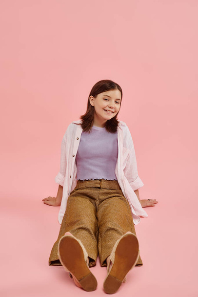 plná délka radostné dospívající dívka v módní ležérní oblečení sedí a usmívá se na kameru na růžové - Fotografie, Obrázek