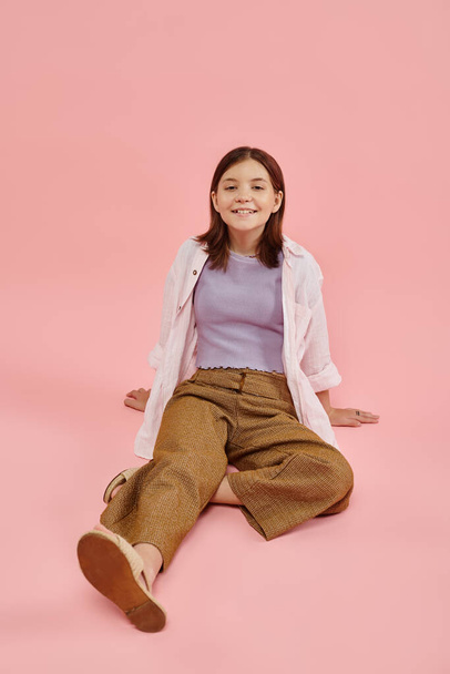longitud completa de alegre adolescente en traje casual de moda sentado y sonriendo a la cámara en rosa - Foto, imagen
