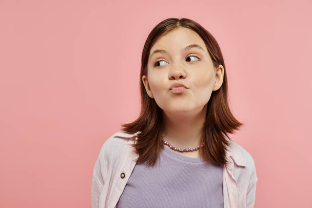 positive and curious teenage girl in trendy clothes pouting lips and looking away on pink backdrop - Photo, Image
