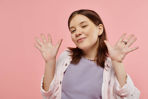 veselý dospívající dívka ve stylové ležérní oblečení mávání rukama a při pohledu na kameru na růžové pozadí - Fotografie, Obrázek