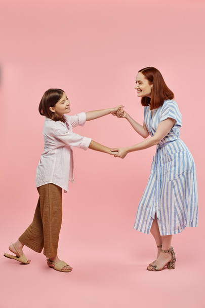 volle Länge der unbeschwerten und trendigen Frau mit frühgeborener Tochter, die Händchen hält und auf rosa tanzt - Foto, Bild