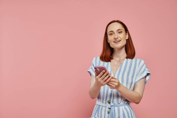 cheerful stylish woman in striped dress holding smartphone and looking at camera on pink backdrop - Photo, Image