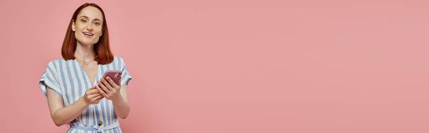 femme gaie et élégante en robe rayée tenant smartphone et regardant la caméra sur rose, bannière - Photo, image