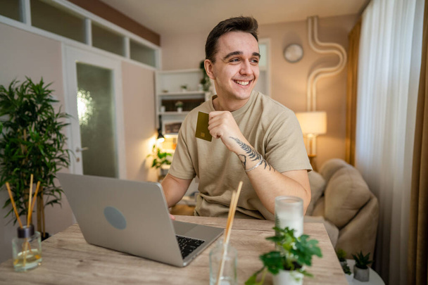 One man adult caucasian male sit at home happy smile at laptop computer hold bank credit card online shopping concept real people copy space e-commerce - Zdjęcie, obraz