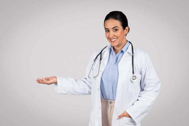 Latin woman doctor in uniform, confidently presenting or showing invisible object on her open palm, facilitating a showcase for any product against grey studio background - Photo, Image