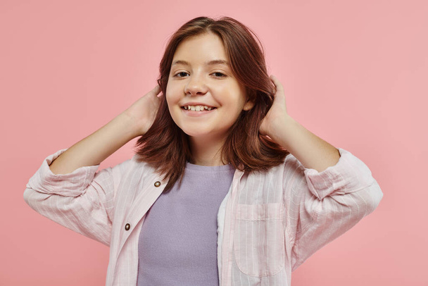 joyful and stylish preteen girl with healthy shiny hair smiling at camera on pink backdrop in studio - Photo, Image