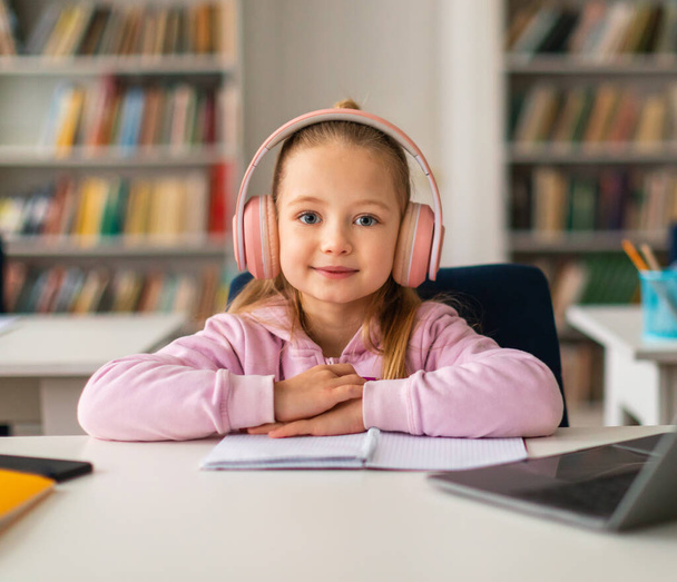 Hezká školačka u stolu, ponořená do online výuky se svým laptopem, s bezdrátovými sluchátky a s úsměvem do kamery, ilustrující lehkomyslný e-learningový moment - Fotografie, Obrázek