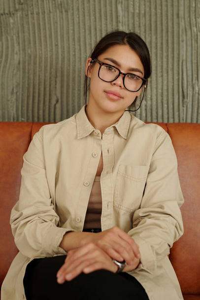 Fiatal barna üzletasszony szemüvegben és bézs ingben ül a kanapén és a kamerát nézi stúdióban vagy pózol fotózás közben - Fotó, kép