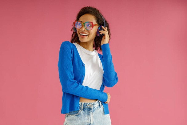 Pretty woman in glasses touching headphones while looking at side on pink studio background - Foto, Imagen