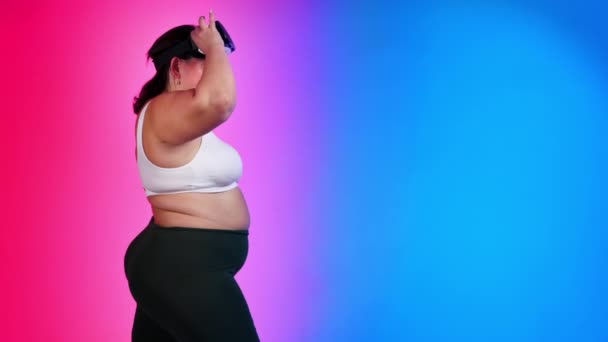 Mujer con sobrepeso en chándal que se pone los auriculares VR mientras posa en un estudio con fondo azul y rojo - Imágenes, Vídeo