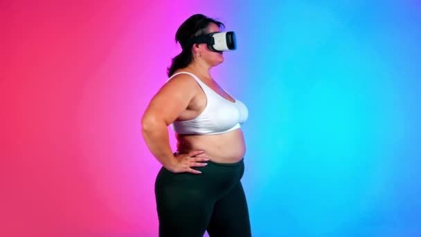 Frau mit Übergewicht im Trainingsanzug und VR-Headset macht Übungen in einem Studio mit blauem und rotem Hintergrund - Filmmaterial, Video
