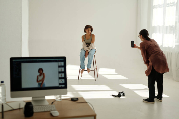 写真のセッション中にスマートフォンでファッションモデルの写真を撮るカジュアルウェアの若い女性写真家 - 写真・画像