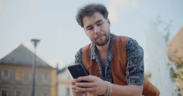 Przystojny młody człowiek SMS-y przez telefon komórkowy w ogrodzie podczas wakacji - Materiał filmowy, wideo