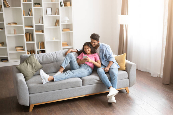 Young indian couple having online shopping, exploring products and offers on their cellphone in a homey ambiance, sitting on sofa, above view, copy space - Photo, Image