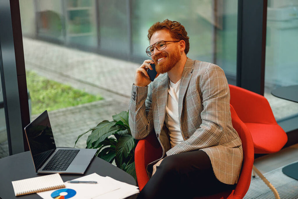 Liikemies puhuu puhelimen asiakkaan kanssa, kun istuu coworking ja työskentelee kannettavan - Valokuva, kuva