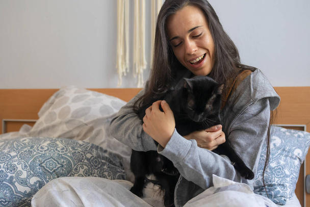 Молодая женщина счастливо улыбается и обнимает свою черную кошку в постели, когда она просыпается утром.. - Фото, изображение