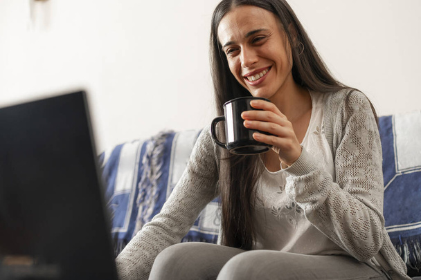 Nainen nauttii kupin teetä, istuu sohvalla kotona, surffatessaan Internetissä kannettavasta tietokoneestaan.. - Valokuva, kuva