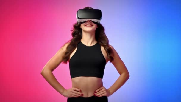 Молода усміхнена жінка в трек-костюмі у віртуальній реальності VR гарнітура дивиться навколо в студії з синім і червоним фоном - Кадри, відео