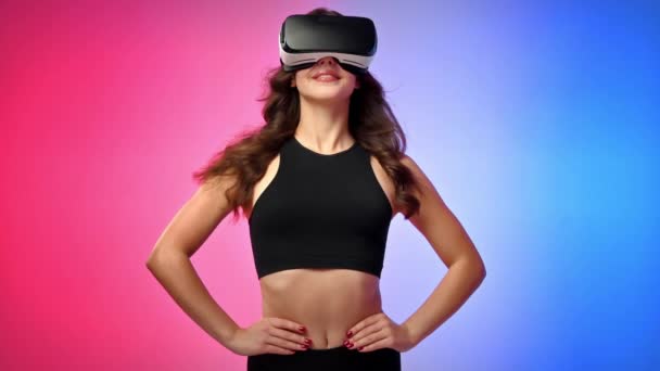 Молода усміхнена жінка в трек-костюмі у віртуальній реальності VR гарнітура позує в студії з синім і червоним фоном - Кадри, відео