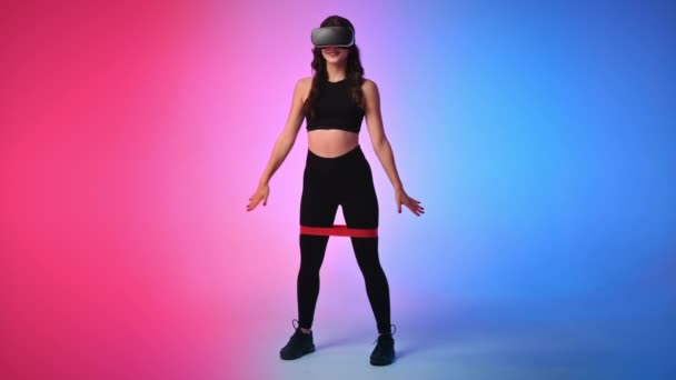Mujer sonriente joven en chándal en realidad virtual Auriculares VR en cuclillas con banda elástica en un estudio con fondo azul y rojo - Metraje, vídeo
