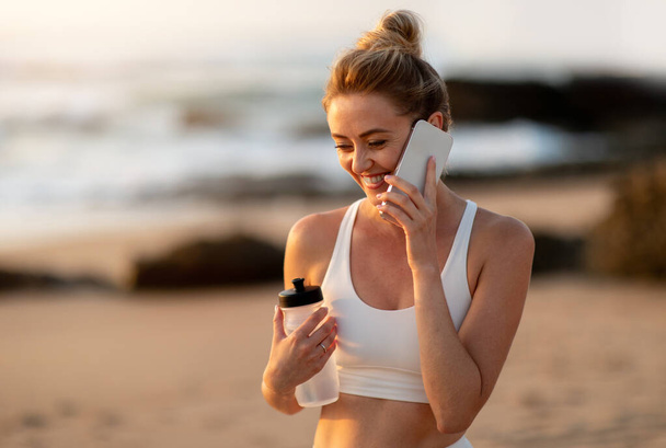 vzrušená běloška, která si během přestávky na venkovním tréninku příjemně popovídá na smartphonu, při západu slunce stojí na mořské pláži, volný prostor - Fotografie, Obrázek