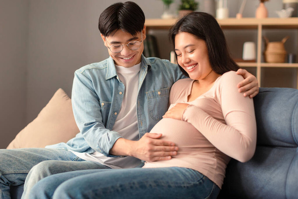 Glückliche Erwartung. Koreanisches junges Paar, das sich über die erste Schwangerschaft freut, sitzt zusammen auf dem heimischen Sofa, der Mann berührt schwangere Frauen am Bauch, umarmt seine Frau im modernen Wohnzimmer. Geburt - Foto, Bild
