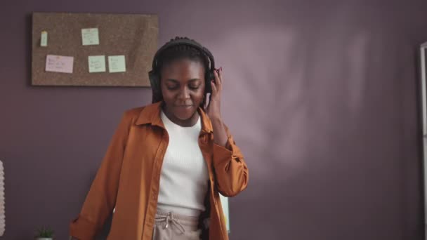 Colpo medio di giovane donna afroamericana vibrante che danza con la musica in cuffia mentre lavora con il marketing di marca da casa - Filmati, video