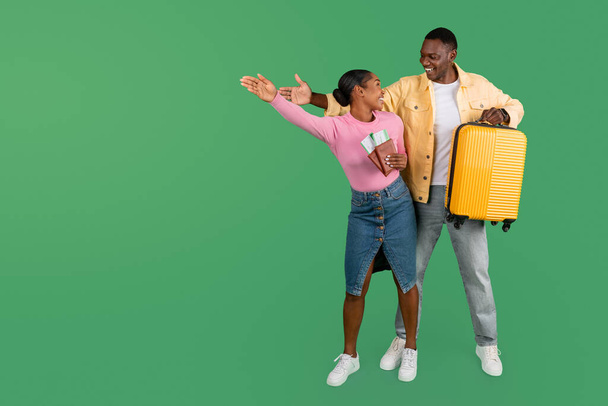 Θετικοί ενθουσιασμένοι νεαροί μαύροι τουρίστες που πηγαίνουν διακοπές μαζί, κρατώντας διαβατήρια και αεροπορικά εισιτήρια, κίτρινη βαλίτσα, δείχνοντας το χώρο αντιγραφής για διαφήμιση, δείχνοντας κατεύθυνση, σε όλο το μήκος - Φωτογραφία, εικόνα