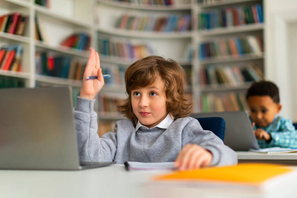 Staranny uczeń, siedzący przed laptopem w tętniącej życiem klasie szkoły podstawowej, podnoszący rękę do walki, wykazujący chęć uczestniczenia i uczenia się podczas lekcji - Zdjęcie, obraz